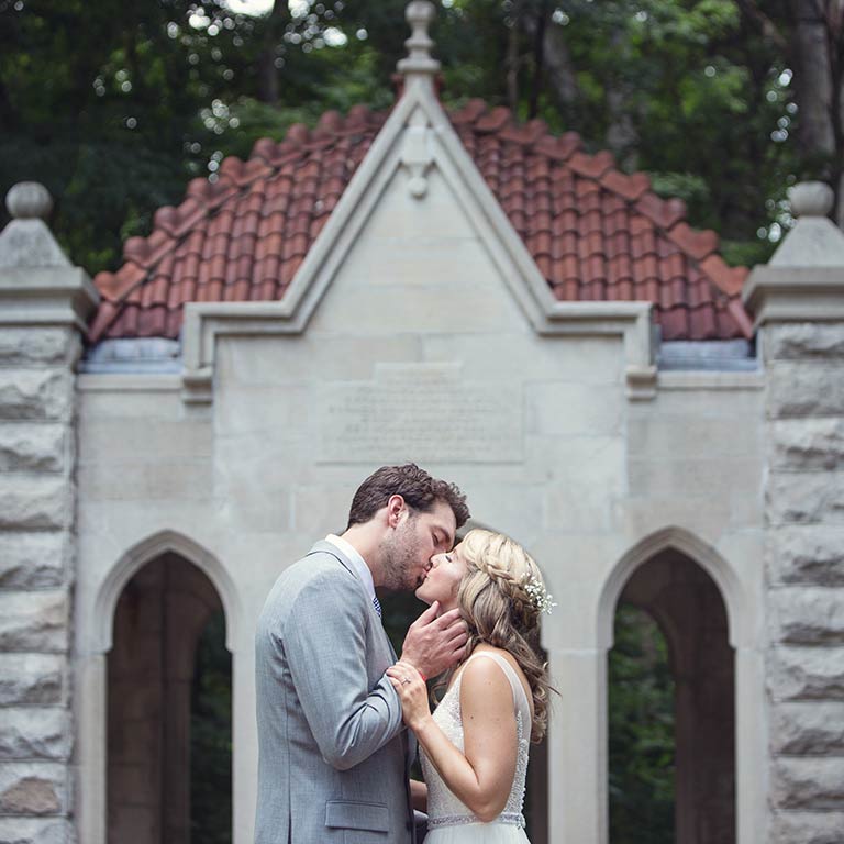 A man and a woman kiss in front of the Rose Well House on their wedding day.